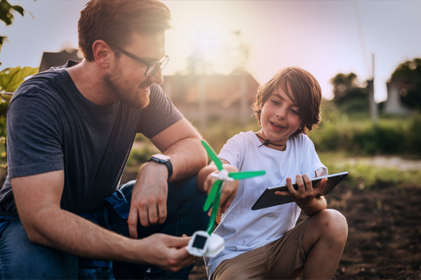 Mężczyzna w okularach i chłopiec są ogrodzie o zachodzie słońca. Wspólnie przyglądają się małej, zabawkowej turbinie wiatrowej, a chłopiec trzyma w ręku tablet.