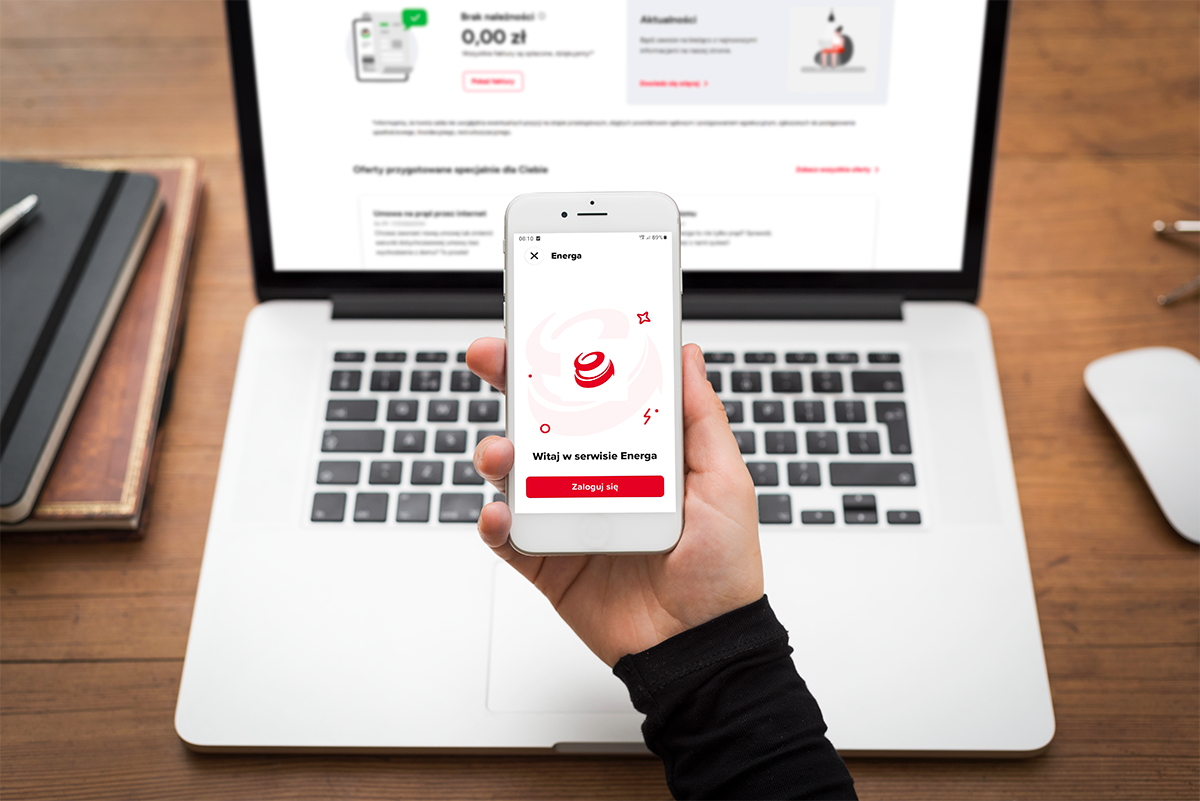 Dłoń trzymająca smartfon z otwartą aplikacją mobilną na tle otwartego laptopa na drewnianym biurku