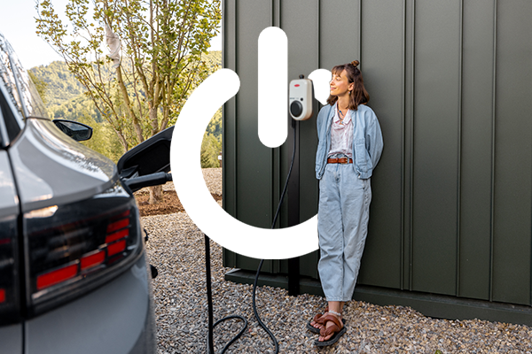 Kobieta stoi przed garażem, na ścianie widoczna stacja ładowania auta elektrycznego. Auto jest podłączone do ladowarki.