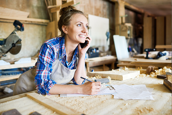 Uśmiechnięta kobieta w roboczym fartuchu rozmawiająca przez telefon i notująca coś przy stole w warsztacie stolarskim