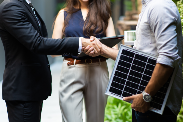 Mężczyzna w garniturze i mężczyzna trzymający panel fotowoltaiczny podają sobie ręce na powitanie. W tle stoi kobieta z tabletem.