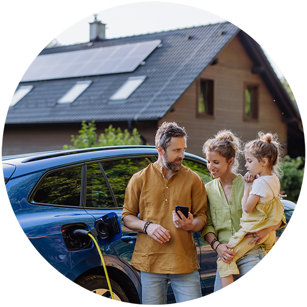 Trzyosobowa rodzina stoi przy ładującym się aucie elektrycznym, w tle dom z instalacją PV na dachu