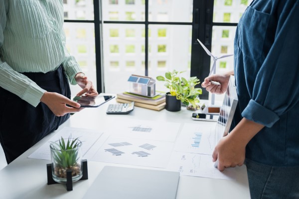 Dwie osoby stojące przy białym biurku, omawiające projekt dotyczący odnawialnych źródeł energii. Na stole widoczne są makiety domu z panelami słonecznymi, turbina wiatrowa, wykresy oraz tablet.