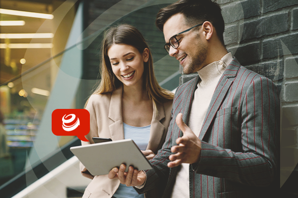 Kobieta i mężczyzna stoją uśmiechnięci w przestrzeni biurowej. Mężczyzna trzyma w ręku tablet i pokazuje ekran tabletu kobiecie. 
