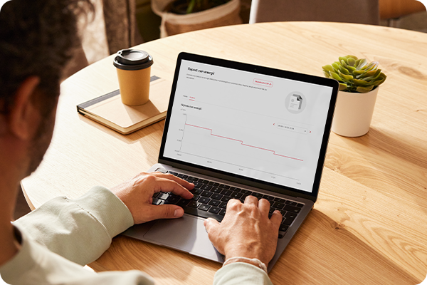 Laptop leży na biurku, na jego ekranie widoczny jest wykres raportu cen energii dla oferty dynamicznej  w ENERGA24. Po lewej stronie leży kubek i notatnik.