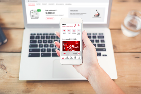 Kobieta trzymająca w dłoni telefon z otwartą aplikacją ORLEN VITAY. W tle laptop z serwisem obsługi ENERGA24.