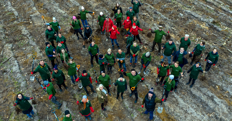 Grupa osób trzymająca sadzonki drzew stoi w kręgu z głowami uniesionymi do góry, ujęcie z drona.
